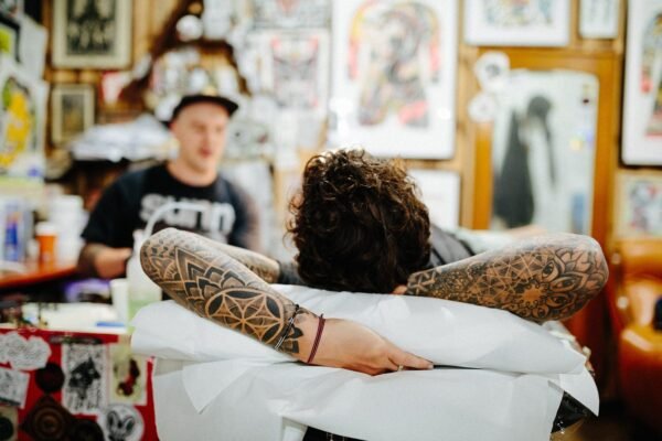 A client with tattooed arms behind his head relaxes at the studio, a tattoo artist sitting behind him